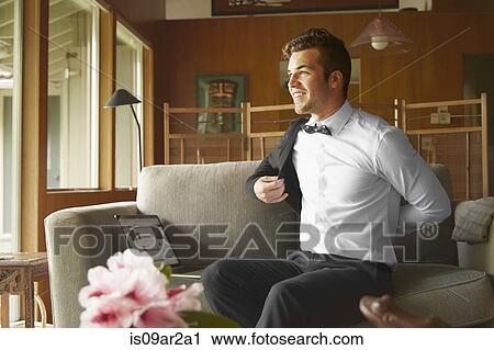 Stock Image - Man in smart clothes sitting on sofa putting on jacket. Fotosearch