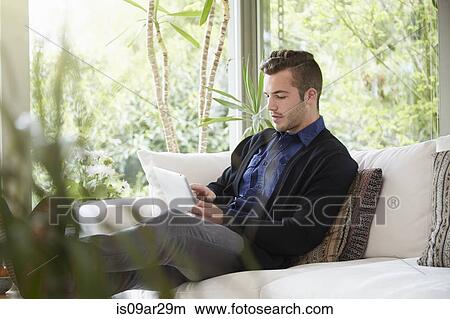 Picture - Man relaxing on sofa with feet up looking at digital tablet. Fotosearch