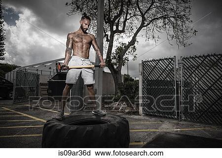 Stock Image - Poised male boxer standing on truck tyre holding sledge hammer in yard. Fotosearch