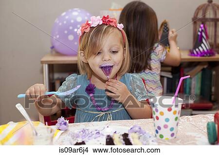 Picture - Portrait of girl sitting at birthday party table eating purple cake with purple tongue. Fotosearch
