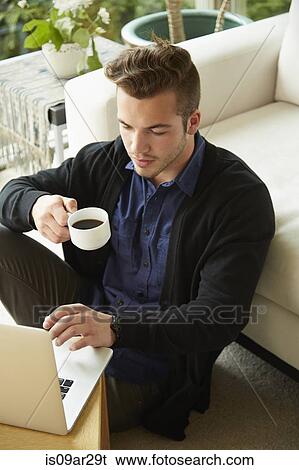 Stock Photograph - Portrait of man sitting on floor at home looking at laptop and drinking cup of coffee. Fotosearch