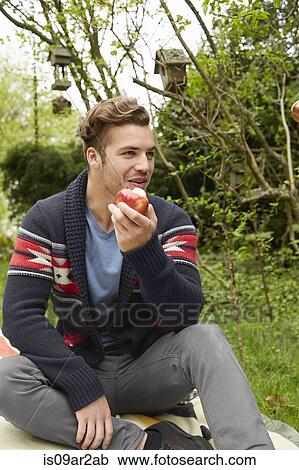 Stock Image - Portrait of smiling man sitting in garden eating an apple. Fotosearch