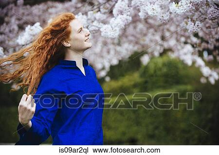 Stock Image - Portrait of young woman with long wavy red hair in spring park. Fotosearch