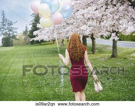 Picture - Rear view of young woman with long wavy red hair and bunch of balloons strolling in spring park. Fotosearch