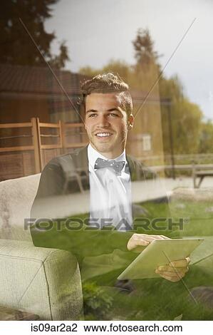 Stock Image - Shot through window of man sitting on sofa using digital tablet. Fotosearch