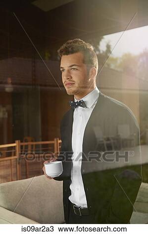 Stock Image - Shot through window of man standing looking out at the view holding cup of coffee. Fotosearch