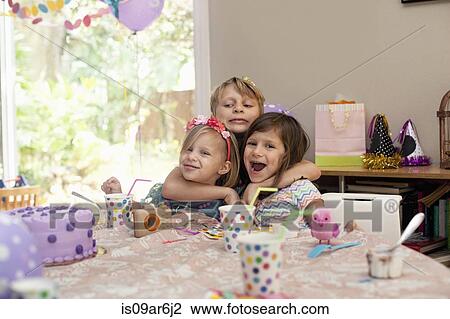 Stock Image - Three children sitting at birthday party table hugging each other. Fotosearch