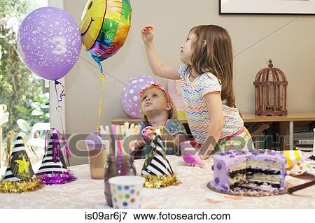 Two girls sitting at birthday party table with cake playing with balloons View Large Photo Image Stock Photo - Two girls sitting at birthday party table with cake playing with balloons. Fotosearch
