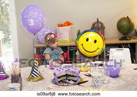 Two girls sitting at birthday party table with cake playing with smiley face balloon View Large Photo Image Stock Photo - Two girls sitting at birthday party table with cake playing with smiley face balloon. Fotosearch