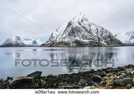 Stock Photograph - "View of snow capped mountain and rocky coastline, Reine, Lofoten, Norway". Fotosearch