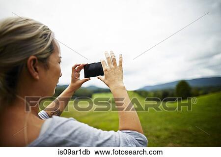 Stock Image - Woman taking photo on smartphone of rural field landscape. Fotosearch