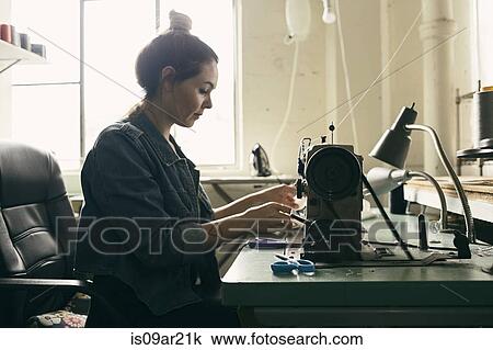 Stock Image - Young female seamstress using sewing machine in fashion studio. Fotosearch
