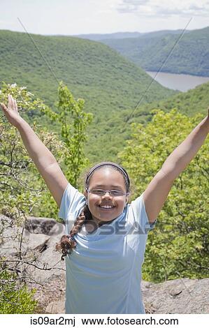 Stock Image - "Young girl at top of hilly landscape, arms raised, smiling". Fotosearch