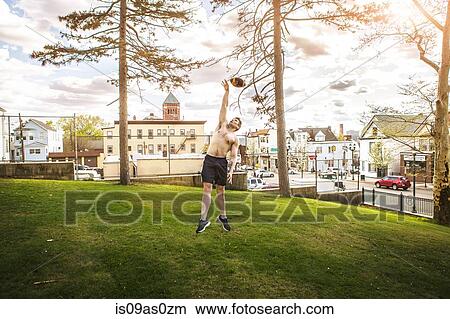 Picture - Bare chested young man jumping to reach American football ball in park. Fotosearch