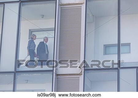 Stock Image - Businessman and woman walking in corridor of glass fronted hotel. Fotosearch