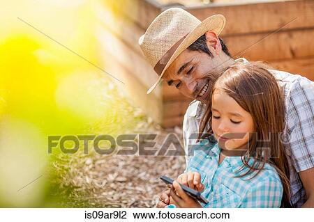 Girl on fathers lap using smartphone in community garden View Large Photo Image Stock Image - Girl on fathers lap using smartphone in community garden. Fotosearch