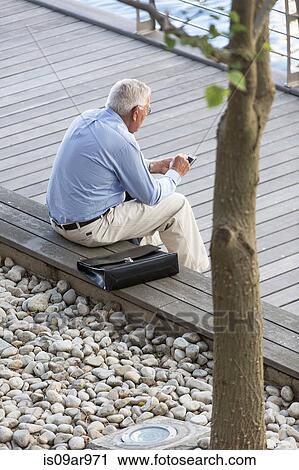 Stock Image - High angle view of senior businessman texting on smartphone on hotel terrace. Fotosearch
