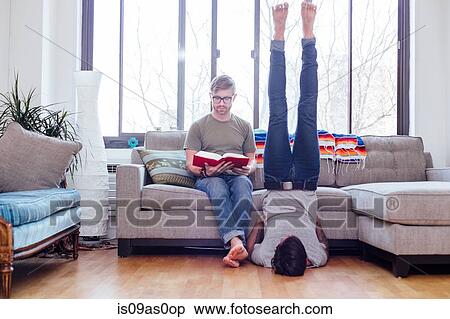 Male couple at home, mid adult male reading whilst partner does shoulder stand View Large Photo Image Stock Photography - Male couple at home, mid adult male reading whilst partner does shoulder stand. Fotosearch