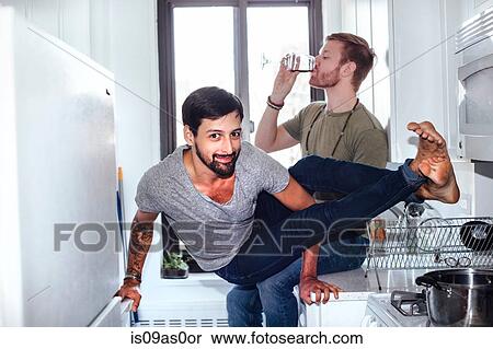 Stock Photography - Male couple at home, mid adult man drinking whilst his partner balances on kitchen appliances. Fotosearch