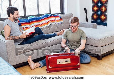 Stock Photograph - Male couple at home, Young man on sofa watching his partner play musical instrument. Fotosearch