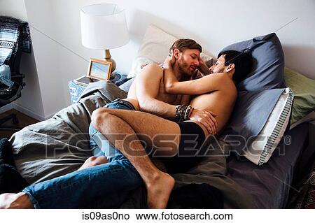 Picture - Male couple, partially dressed, lying on bed, embracing. Fotosearch