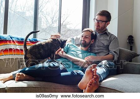 Stock Photo - Male couple relaxing on sofa together with cat. Fotosearch