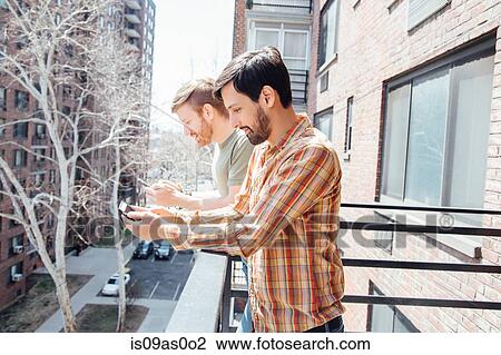 Stock Image - Male couple standing on balcony, looking at smartphones. Fotosearch