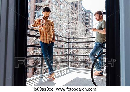 Stock Image - Male couple standing on balcony, standing away from each other, drinking wine and looking at smartphone. Fotosearch