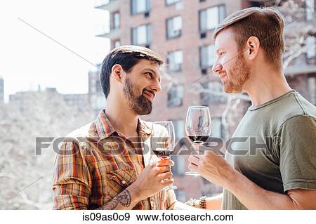 Stock Photo - Male couple standing on balcony, holding wine glasses, face to face. Fotosearch