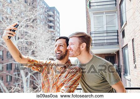 Stock Image - Male couple standing on balcony, taking self portrait, using smartphone. Fotosearch