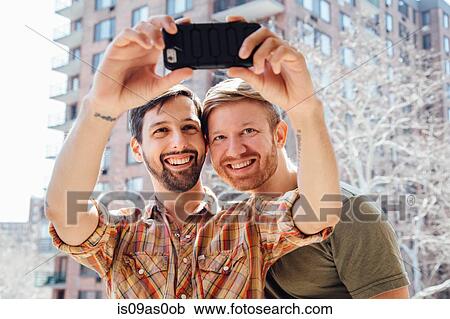 Stock Image - Male couple standing on balcony, taking self portrait, using smartphone. Fotosearch