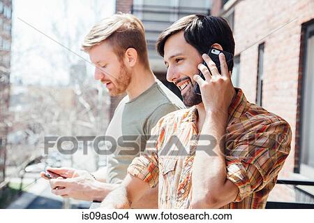 Picture - Male couple standing on balcony, using smartphones. Fotosearch