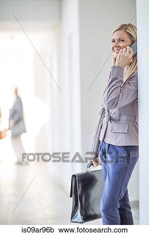 Stock Image - Mid adult businesswoman chatting on smartphone in hotel corridor. Fotosearch