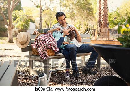 Stock Image - Mid adult man with hand on daughters forehead on community garden bench. Fotosearch
