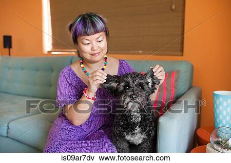 Stock Photo - Mid adult woman sitting on sofa with dog, holding dog's ears. Fotosearch