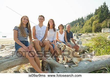 Stock Photograph - Portrait of mature parents, three daughters with dog sitting on beached tree. Fotosearch