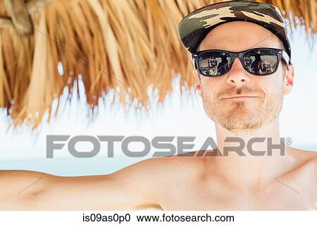 Stock Image - Portrait of mid adult man under cabana, wearing cap and sunglasses, Miami Beach, Florida, USA. Fotosearch