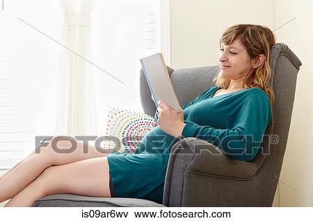 Stock Photo - Pregnant woman reading book on living room armchair. Fotosearch