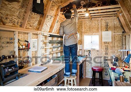 Stock Image - Rear view of leather craftsman searching workshop shelf. Fotosearch