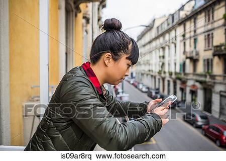 Stock Photography - Side view of woman leaning against balcony using smartphone, Milan, Italy. Fotosearch