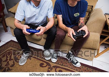 Two young men sitting on sofa, playing video game, low section View Large Photo Image Stock Image - Two young men sitting on sofa, playing video game, low section. Fotosearch