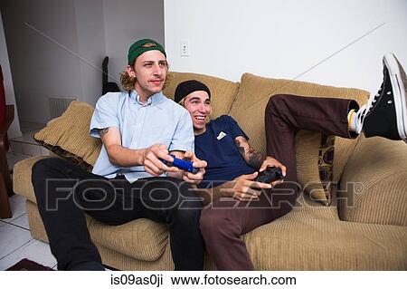 Two young men sitting on sofa, playing video game View Large Photo Image Stock Image - Two young men sitting on sofa, playing video game. Fotosearch
