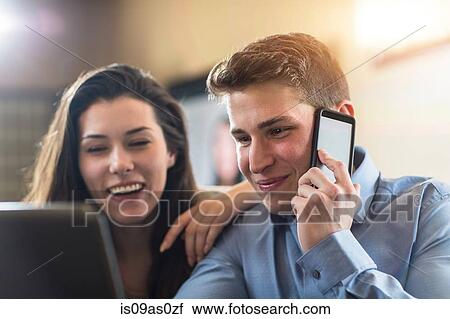 Stock Image - Young couple browsing laptop and talking on smartphone at home. Fotosearch