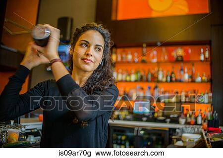 Stock Image - Young female bartender shaking cocktail shaker in cocktail bar. Fotosearch