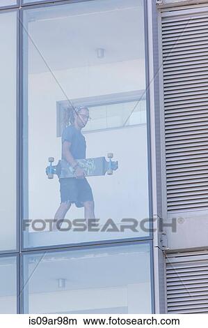 Picture - Young male skateboarder walking in corridor of glass fronted hotel. Fotosearch