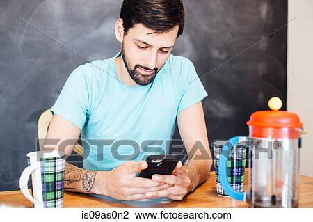Stock Image - Young man sitting at table, having coffee, looking at smartphone. Fotosearch