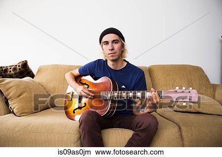 Young man sitting on sofa, playing guitar View Large Photo Image Picture - Young man sitting on sofa, playing guitar. Fotosearch