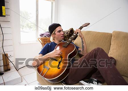 Young man sitting on sofa, playing guitar View Large Photo Image Stock Image - Young man sitting on sofa, playing guitar. Fotosearch