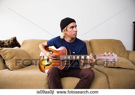 Young man sitting on sofa, playing guitar View Large Photo Image Stock Image - Young man sitting on sofa, playing guitar. Fotosearch