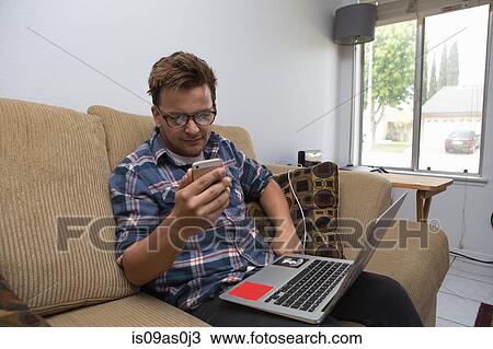 Young man sitting on sofa, using laptop and looking at smartphone View Large Photo Image Stock Image - Young man sitting on sofa, using laptop and looking at smartphone. Fotosearch